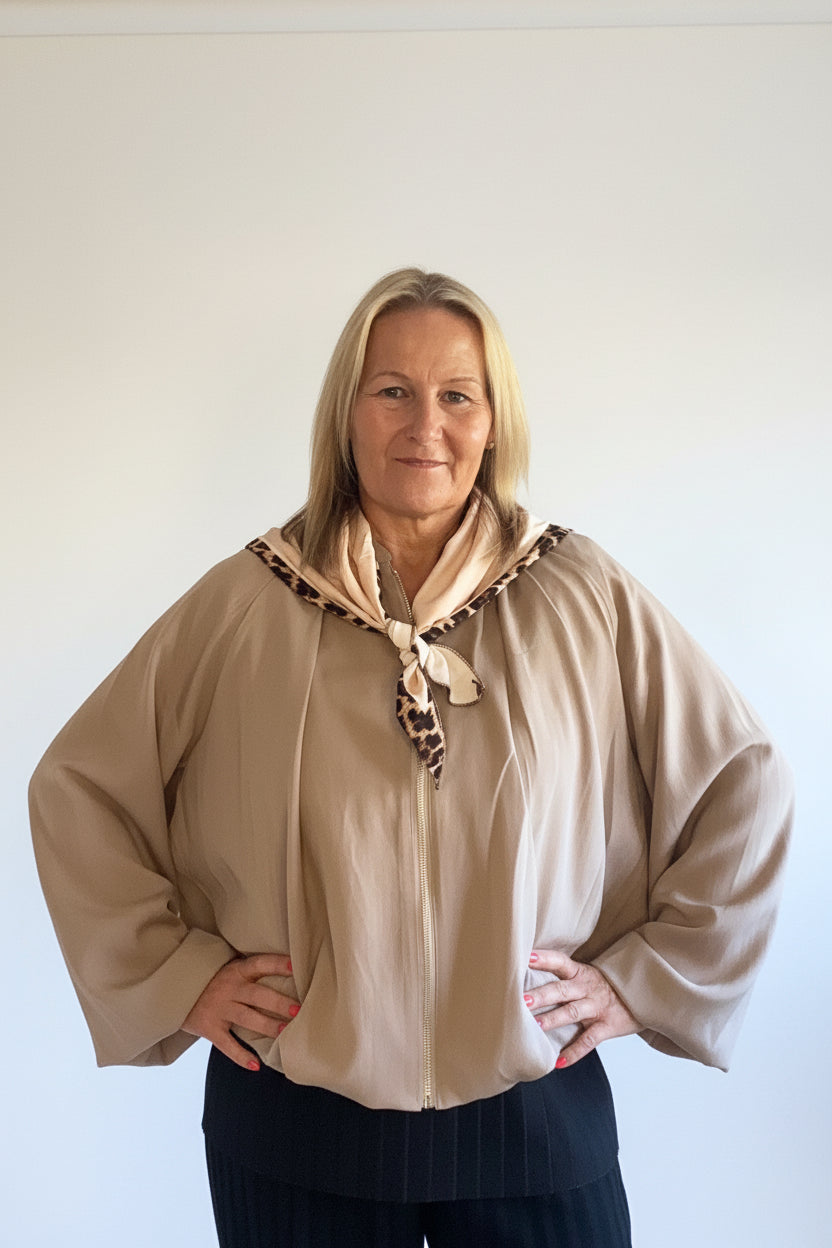 A woman wearing a beige long-sleeve blouse with a patterned tie-neck detail and dark pinstriped trousers, standing in a living room with gray walls and a sofa in the background.