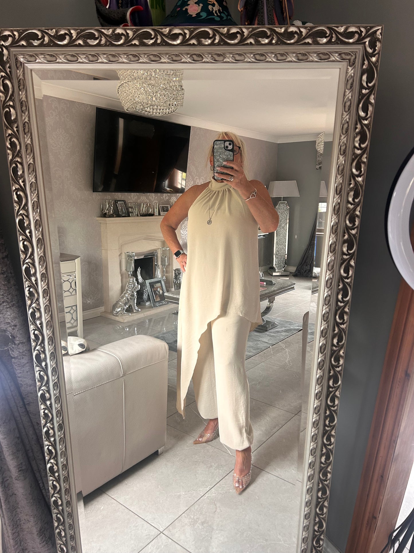 Person wearing a long, light beige wearing a suit standing in front of a decorative mirror.