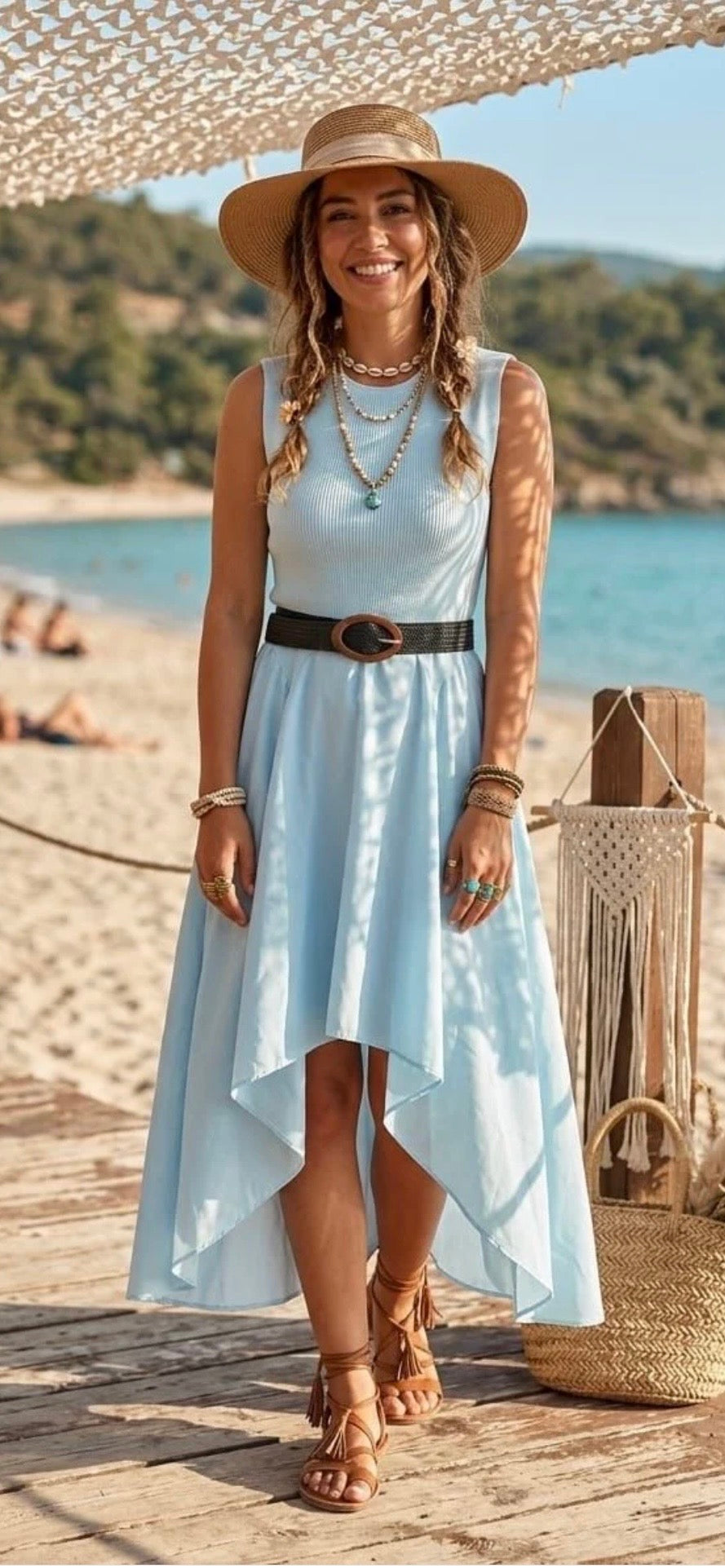 Woman in a light blue dress and straw hat standing on a beach with a scenic background.