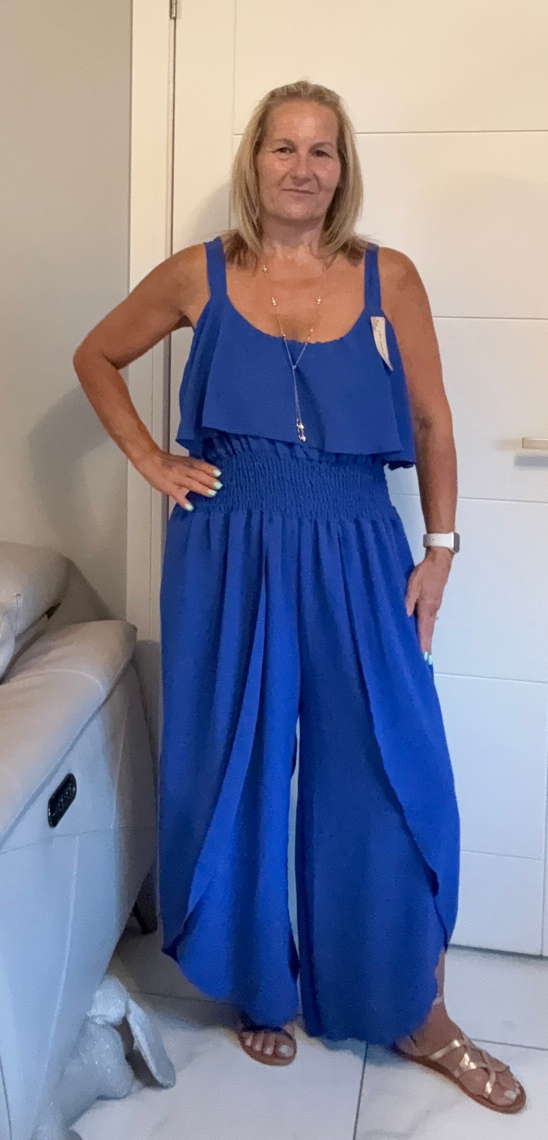 A woman modeling a royal blue sleeveless jumpsuit with a smocked waist and wide legs, standing in a room with white paneled walls.