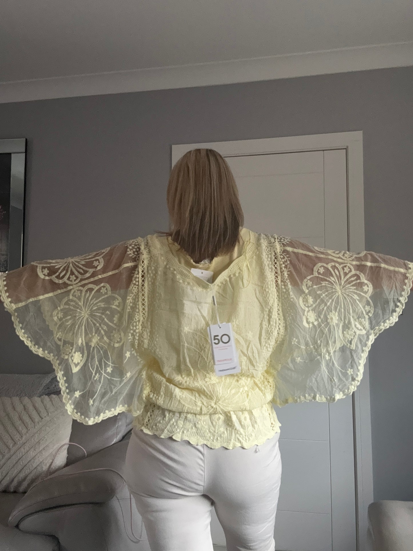 A pale yellow lace-trimmed blouse with dramatic ruffled sleeves, worn by a model in white pants in an indoor room setting.