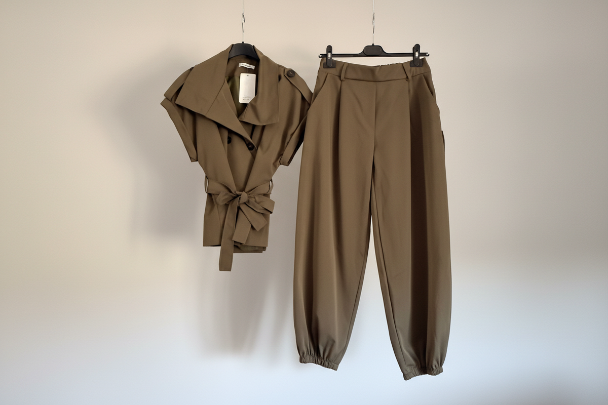 A matching two-piece outfit consisting of a short-sleeve belted wrap-style top and high-waisted tapered pants, both in a warm taupe-brown color, hanging on black hangers against a plain off-white wall.