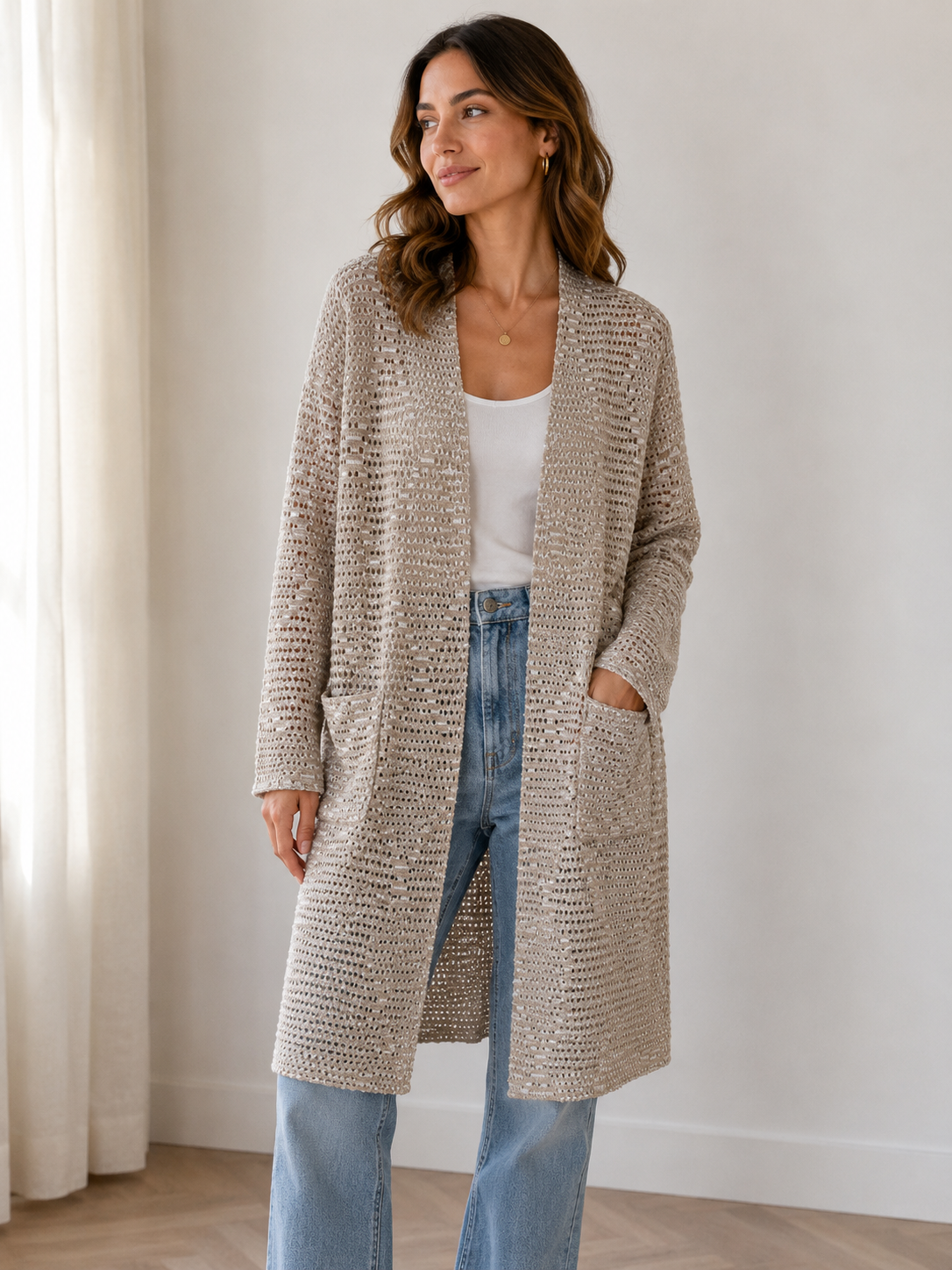 A woman wearing a long, open-front crochet cardigan in a warm cream or beige color, styled over a white tank top and light blue jeans, standing in a brightly lit room with a white curtain