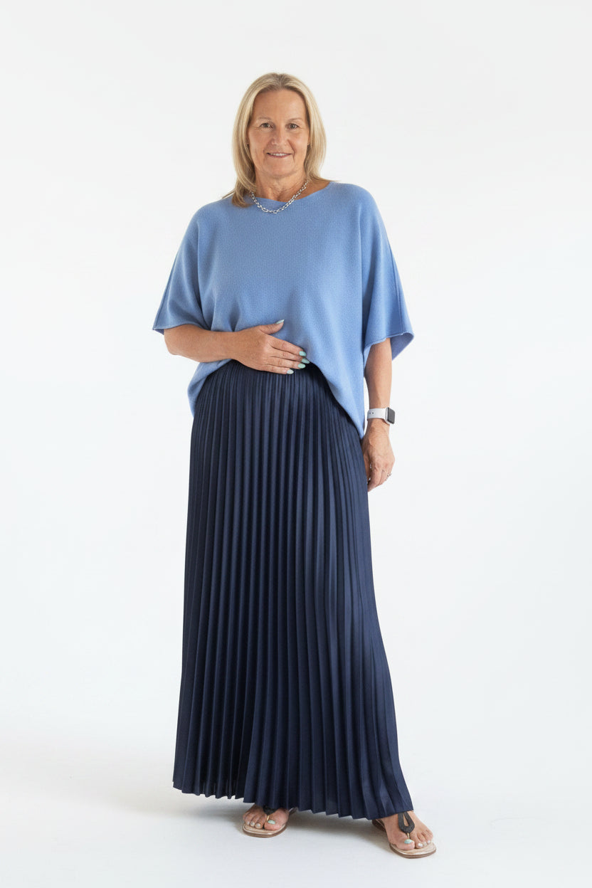 A woman modeling a light blue short-sleeve top and a long navy pleated maxi skirt, standing in a tiled indoor space.