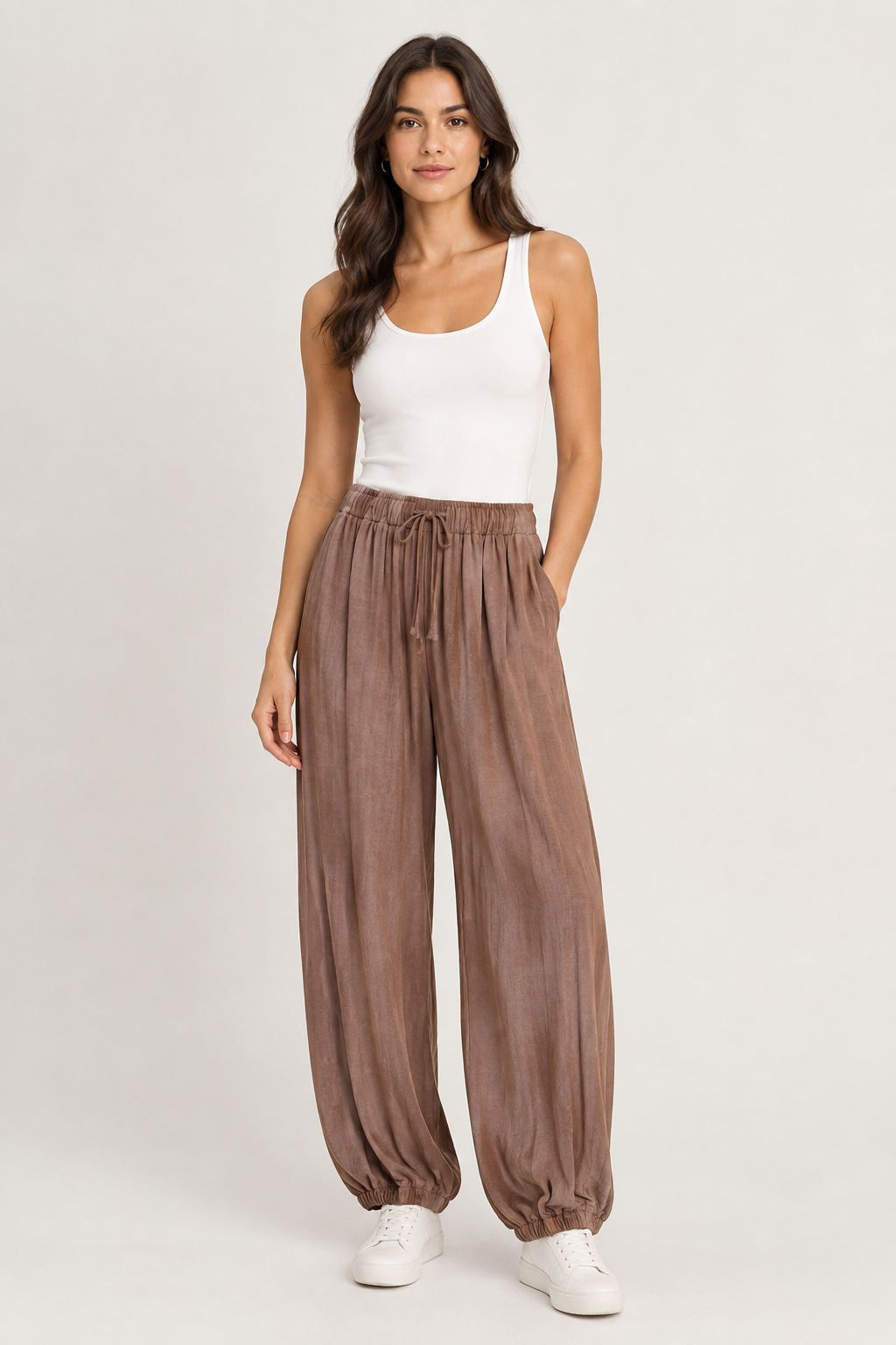 A woman modeling high-waisted, wide-leg brown pants with a drawstring waist, paired with a white tank top and white sneakers, against a light gray studio background.