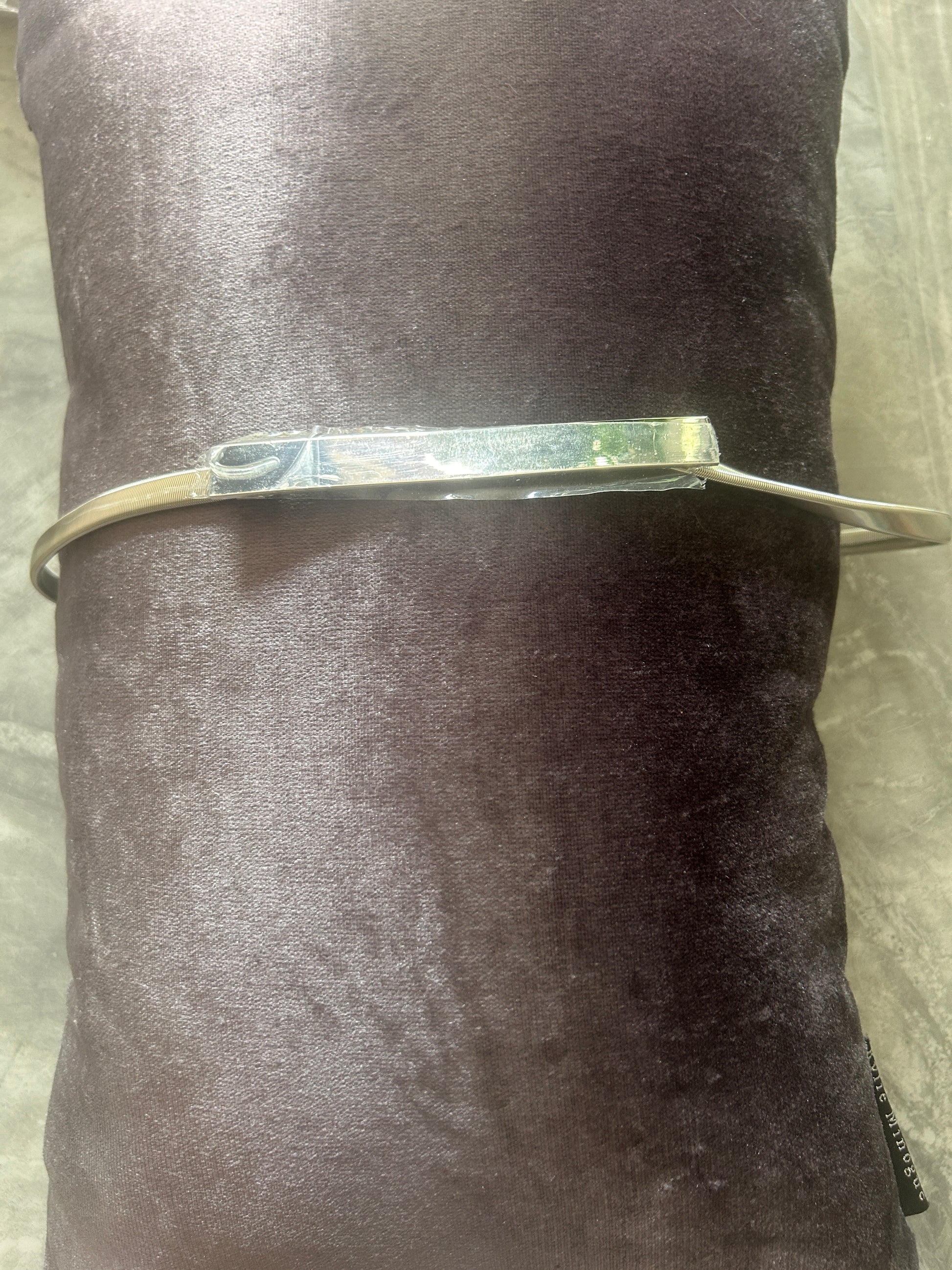 A silver-toned metal bangle belt with a rectangular link and a small loop detail, shown wrapped around a dark brown velvet cylindrical prop on a light gray textured surface.