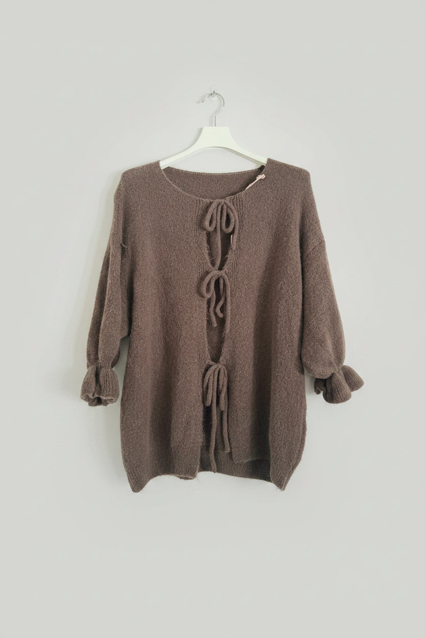 A brown fuzzy knit sweater with three tie-front closures, hanging on a white hanger against a white paneled wall.