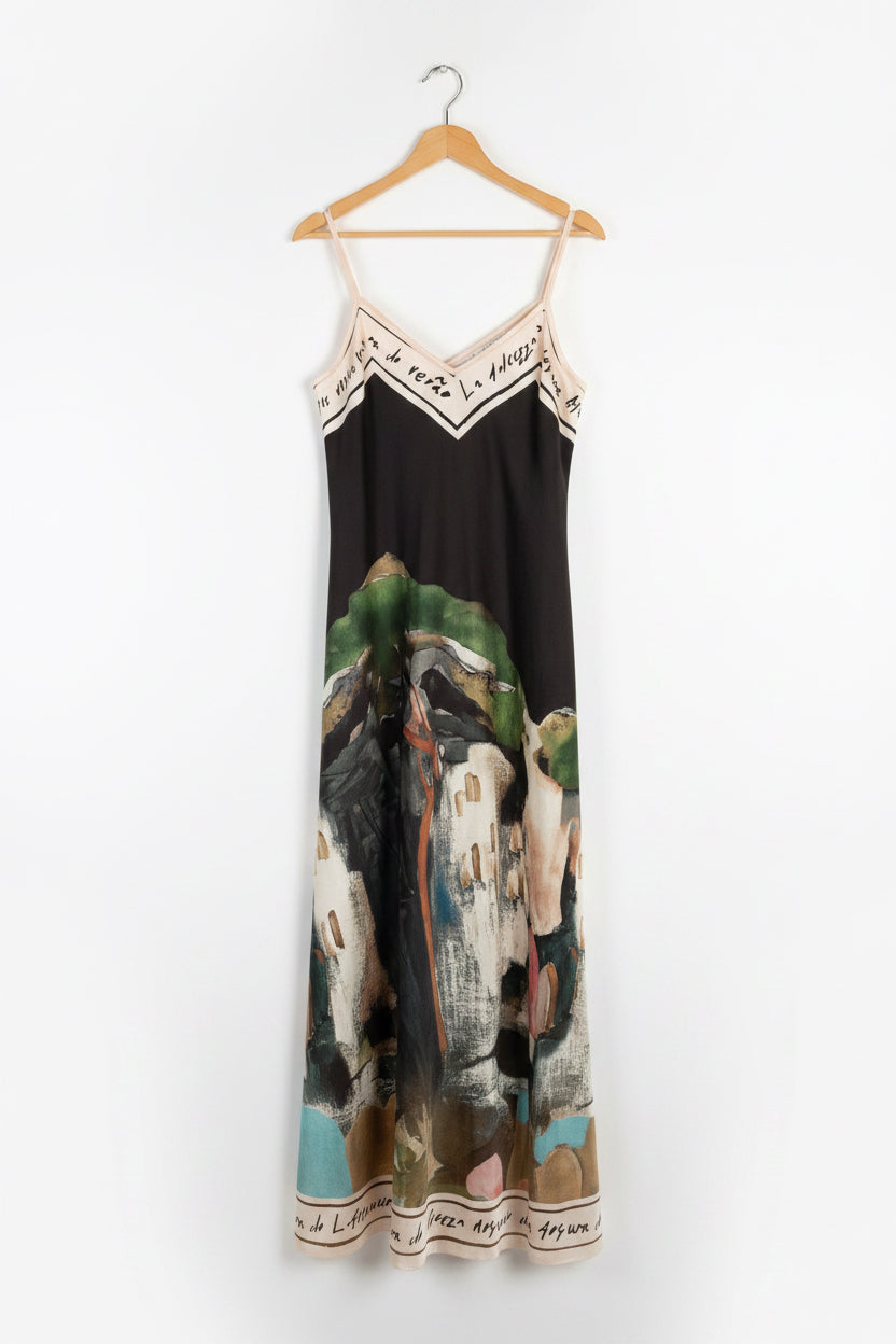 A sleeveless V-neck slip dress with thin straps, featuring a dark brown upper section and a colorful artistic print of a woman on horseback across the lower half. The dress is hanging on a white door. 