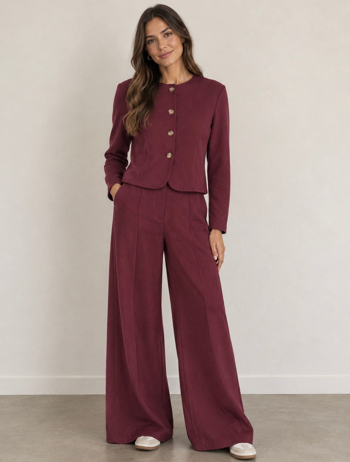 A woman modeling a matching burgundy two-piece linen suit with a cropped jacket and wide-leg trousers, standing against a neutral off-white wall.