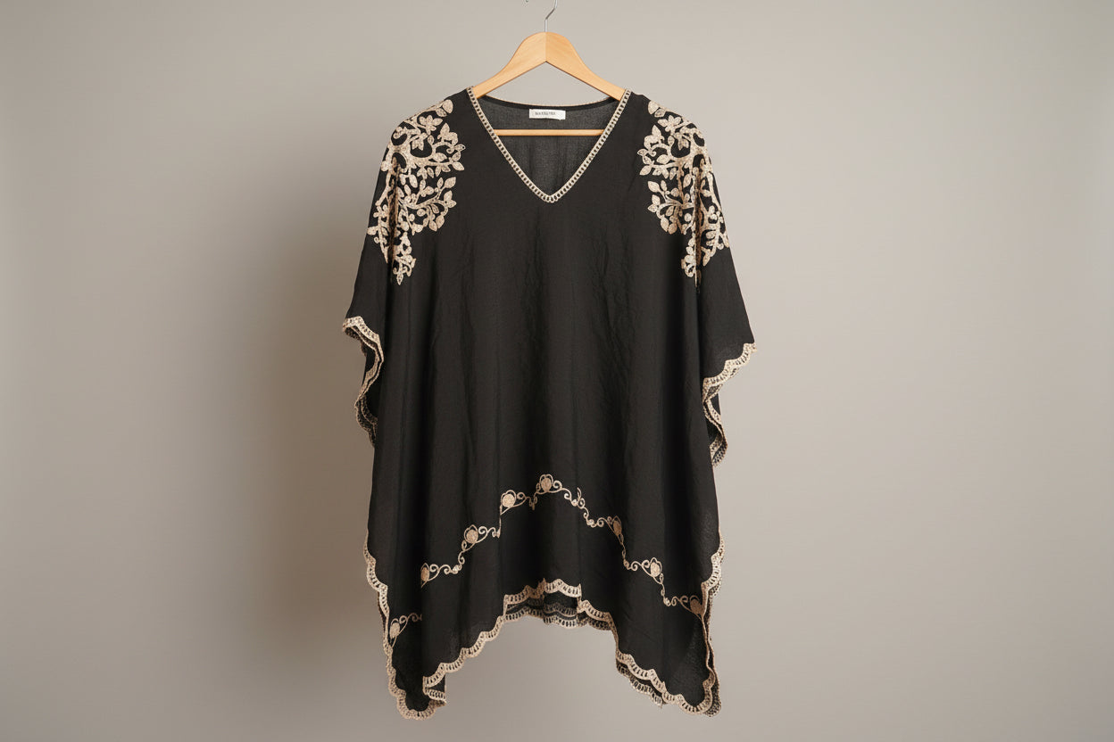 A black V-neck tunic top with beige floral embroidery and scalloped lace trim, hanging on a white wooden hanger against a white paneled wall.