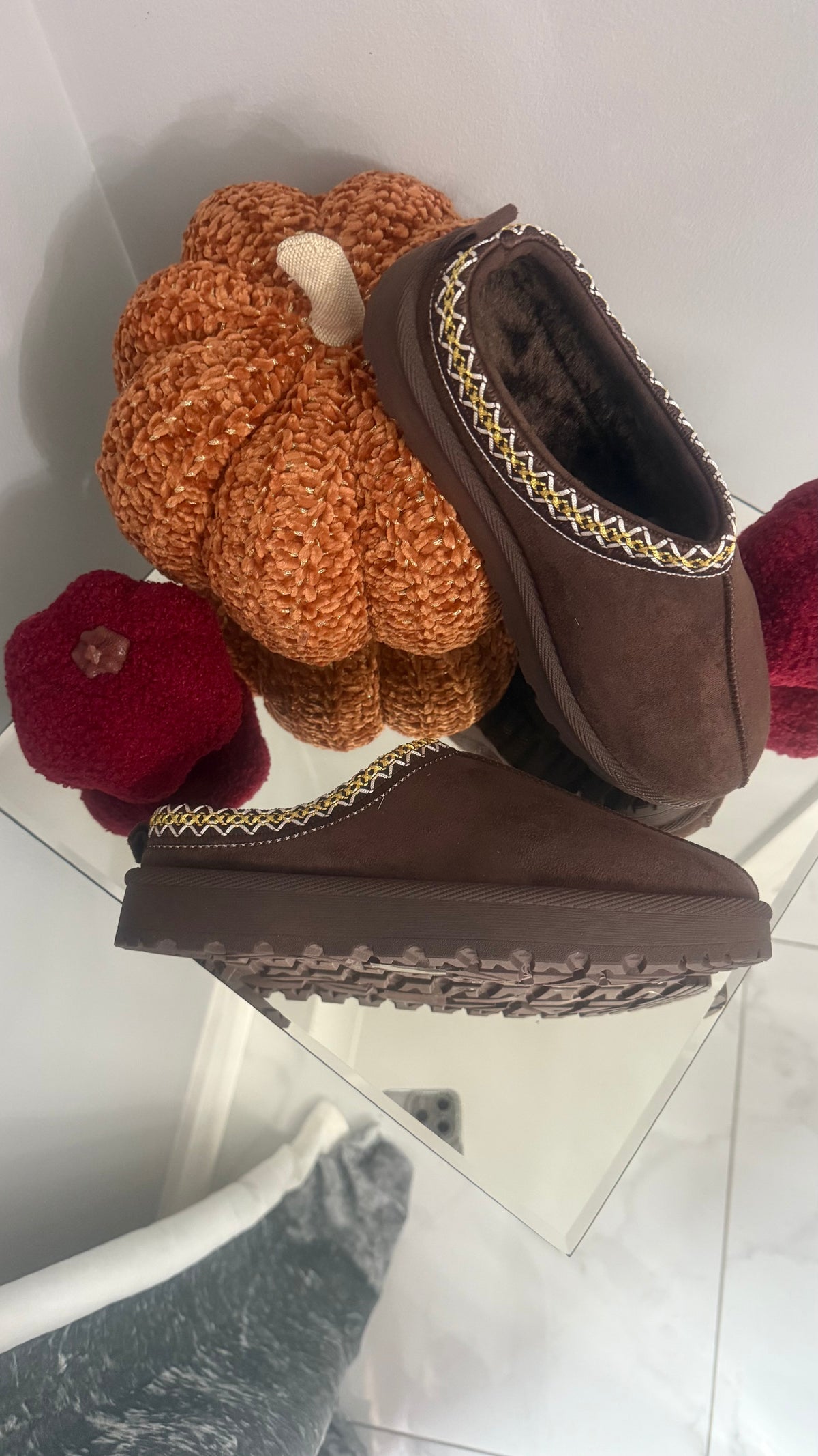 Brown slipper with a knitted pumpkin on top, placed on a reflective surface.