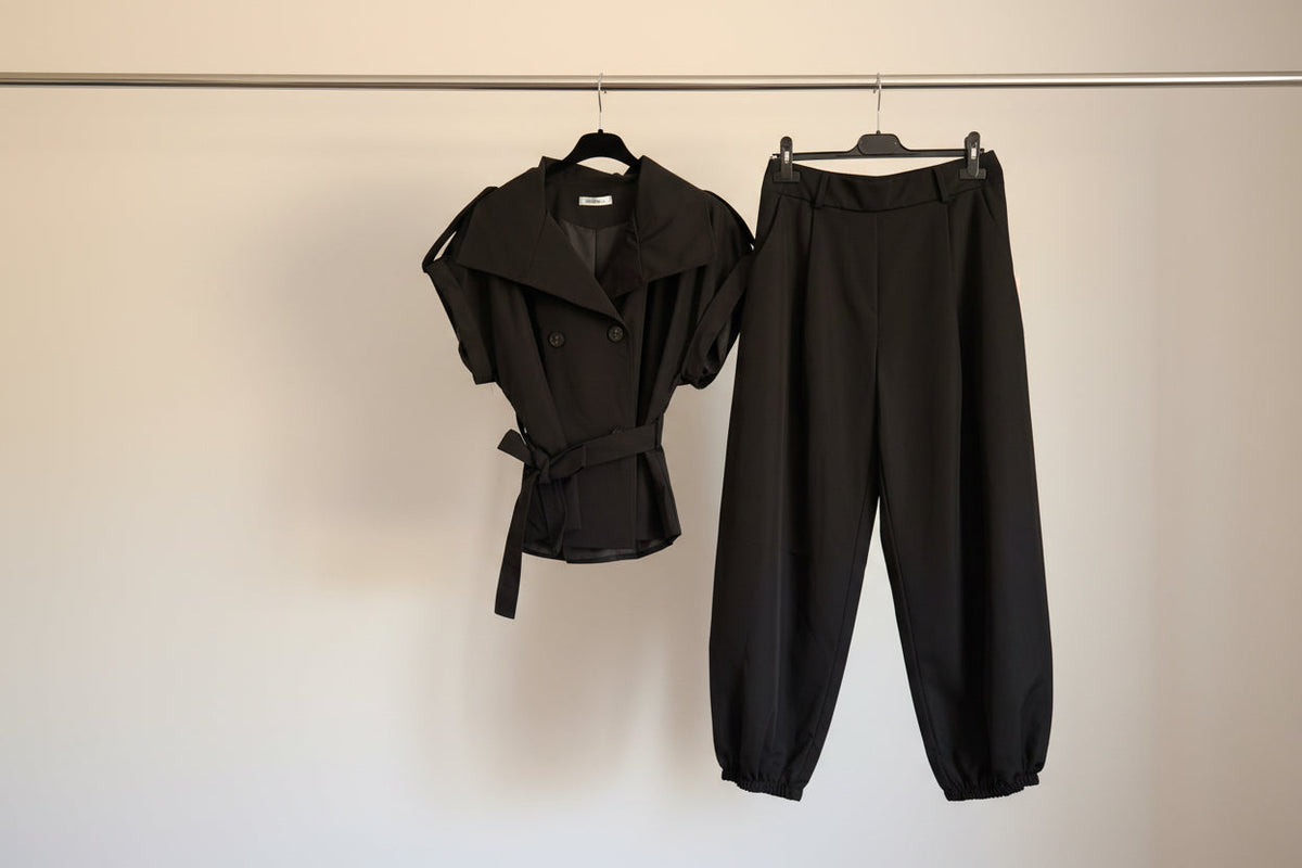 A black short-sleeve belted wrap-style top and black wide-leg cropped pants hanging on black hangers from a metal rail against a plain off-white wall.