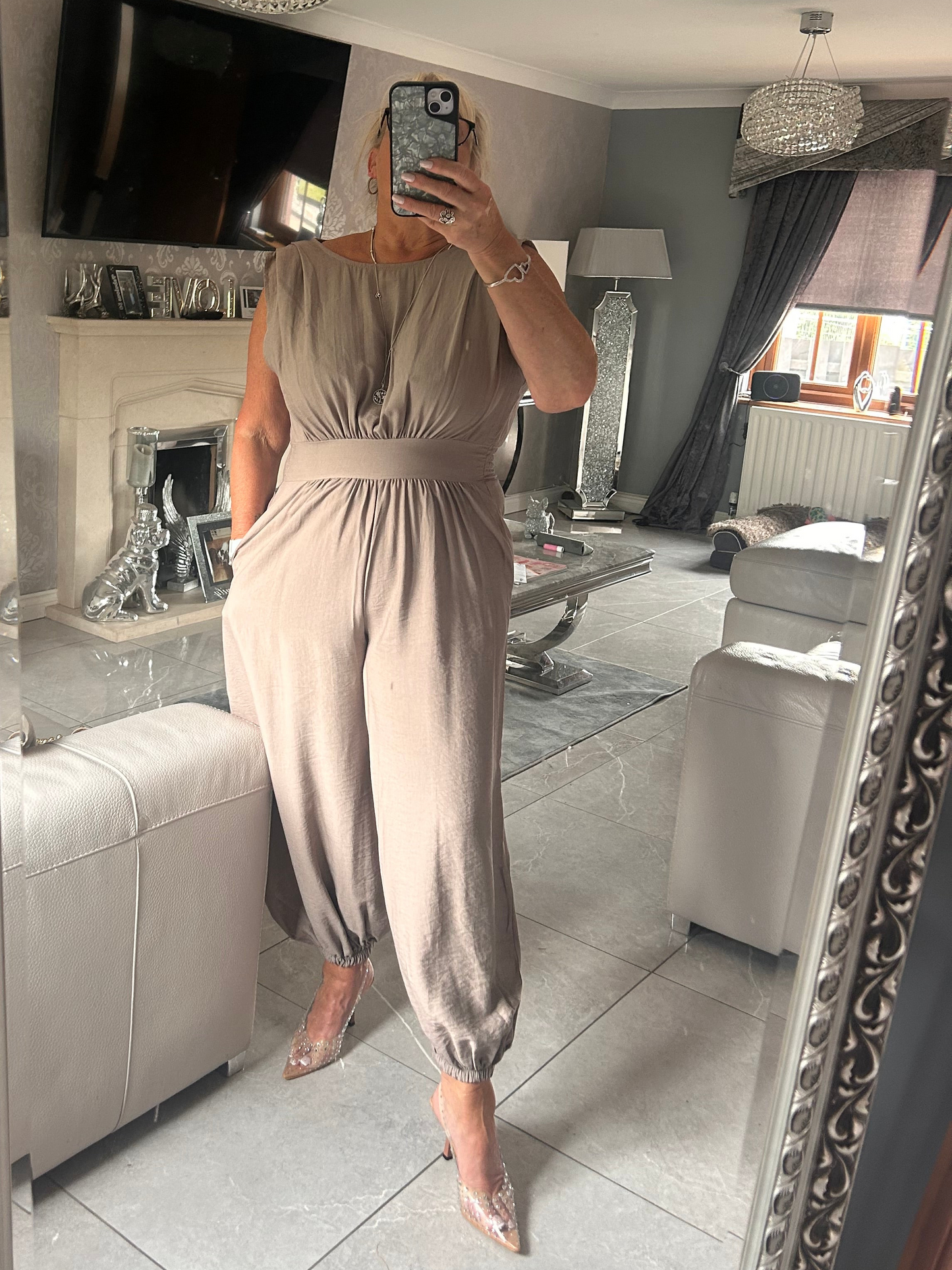 Person wearing a beige jumpsuit taking a mirror selfie in a living room.